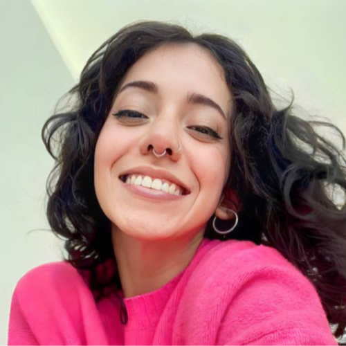 Modello di OnlyFans in Italia, sorridente con capelli ricci e indossando un maglione rosa, posando per un selfie. Contenuto pornografico.